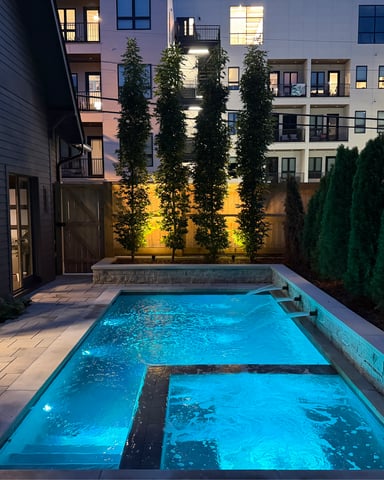 Illuminated swimming pool in courtyard surrounded by ivy-covered walls, evergreens, and multi-story residential buildings at dusk.