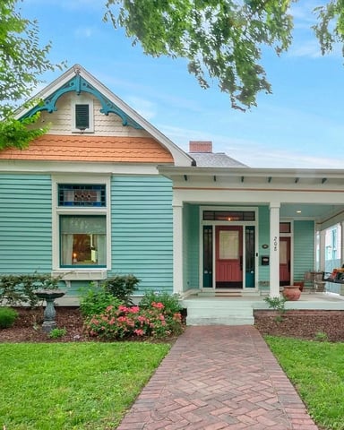Turquoise cottage with red door, covered porch, brick walkway, flowering shrubs, and white picket fence