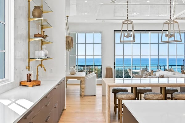 Modern kitchen and dining area with ocean view through floor-to-ceiling windows, brass shelving, pendant lights, and wooden bar seating.