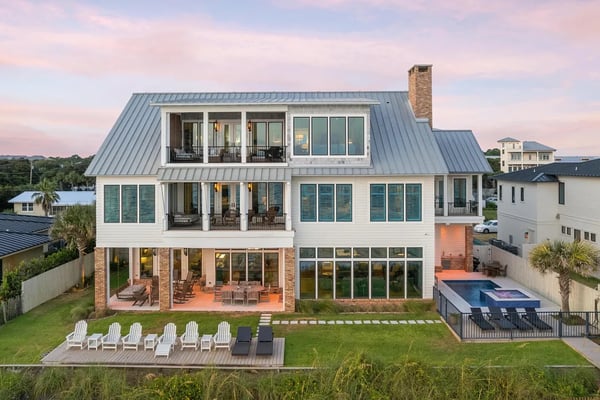 Modern three-story coastal home with metal roof, brick chimney, multiple decks, and backyard pool at sunset.