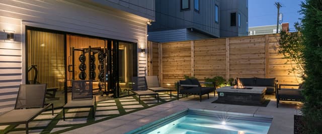 Modern backyard with illuminated pool, lounge chairs, fire pits, wooden fence, and contemporary house at dusk