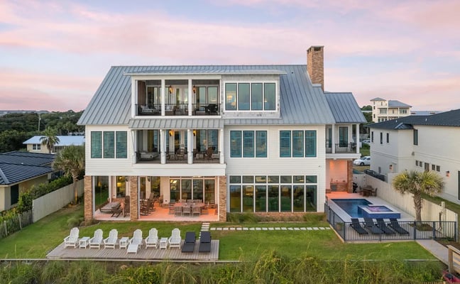 Modern three-story coastal home with metal roof, brick chimney, multiple decks, and backyard pool at sunset.