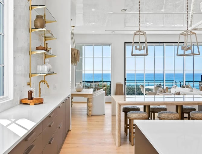 Modern kitchen and dining area with ocean view, brass shelving, pendant lights, and bar seating overlooking blue water.
